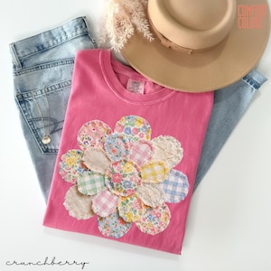 May include: A pink t-shirt with a floral applique made of various patterned fabrics. The shirt is paired with light-wash jeans, a tan hat, and a necklace. The text "COMFORT COLORS" is visible on the hat.
