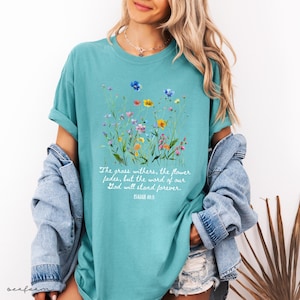 May include: Teal-colored t-shirt featuring a watercolor floral design with various colorful wildflowers. The shirt includes the text "The grass withers, the flower fades, but the word of our God will stand forever. ISAIAH 40:8."