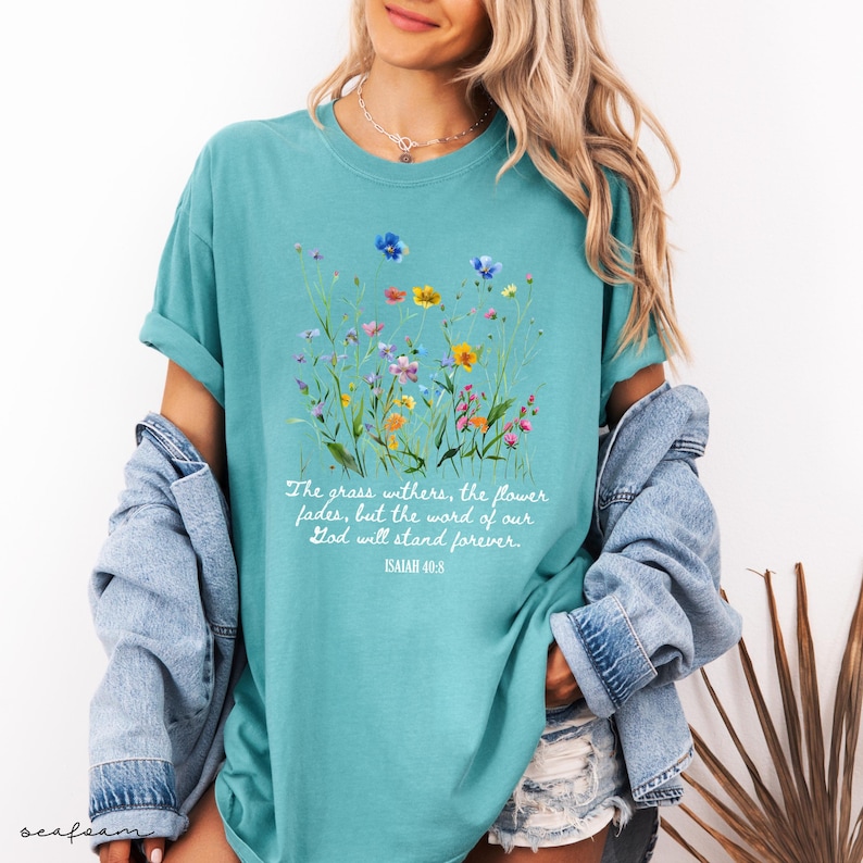 May include: Teal-colored t-shirt featuring a watercolor floral design with various colorful wildflowers. The shirt includes the text "The grass withers, the flower fades, but the word of our God will stand forever. ISAIAH 40:8."