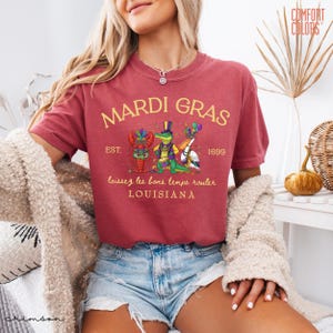 May include: Crimson-colored t-shirt with "MARDI GRAS" in gold lettering. The shirt features a graphic of a crawfish, alligator, and pelican in festive attire, with the text "laissez les bons temps rouler" and "LOUISIANA".