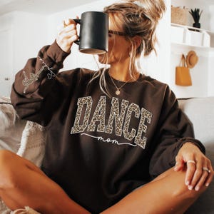 May include: A dark brown sweatshirt with the text "DANCE mom" in a leopard print design. The sweatshirt has a crew neck and long sleeves. A small dancer graphic is on the left sleeve. The person is holding a black mug.