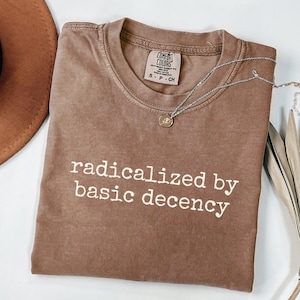 May include: A brown t-shirt with the text "radicalized by basic decency" printed in white on the front.