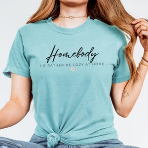 May include: A turquoise blue t-shirt with the text "Homebody I'd rather be cozy at home." in black lettering and a small white daisy graphic.