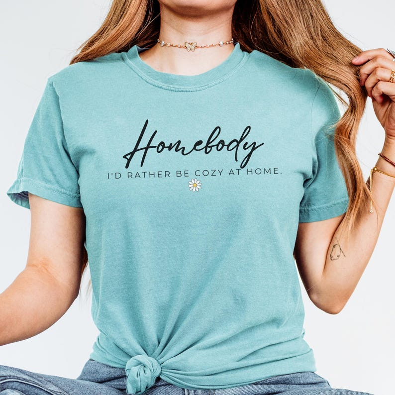 May include: A turquoise blue t-shirt with the text "Homebody I'd rather be cozy at home." in black lettering and a small white daisy graphic.