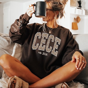 May include: A brown sweatshirt with the name "CECE" in leopard print lettering and the text "est. 2026". The sleeve has the names "Olivia", "Quinn", and "Ethan". A person is holding a black mug.