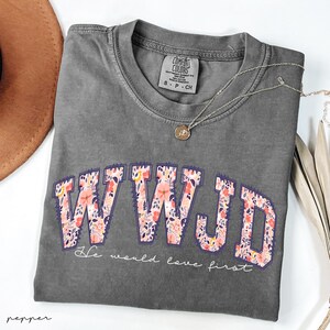 May include: A grey Comfort Colors t-shirt featuring the letters "WWJD" in a floral pattern. The text "He would love first" is below. A silver necklace with a pendant is draped on the shirt.