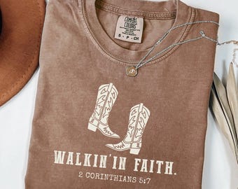 Könnte beinhalten: Ein braunes T-Shirt mit ausgewaschenem Look, das ein Motiv mit zwei Cowboystiefeln und dem Text "WALKIN' IN FAITH. 2 CORINTHIANS 5:7" zeigt.