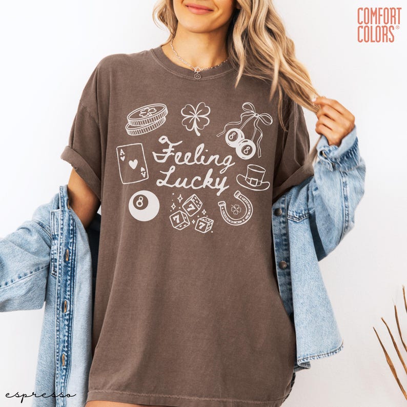 Może przedstawiać: Brązowy t-shirt z napisem "Feeling Lucky" i r&oacute;żnymi białymi symbolami szczęścia. Projekt zawiera czterolistną koniczynę, kości, podkowę i cylinder. Marka Comfort Colors.
