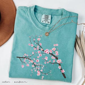 May include: Seafoam green t-shirt with a pink cherry blossom branch graphic. The shirt has a crew neck and short sleeves. The text "COMFORT COLORS" is visible on the tag.