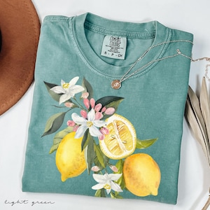 May include: Light green t-shirt with a watercolor lemon and floral design. The shirt features a crew neck and short sleeves. A silver necklace with a small pendant is draped across the shirt. The words "light green" are written at the bottom.