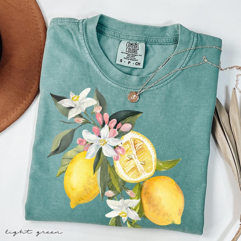 May include: Light green t-shirt with a watercolor lemon and floral design. The shirt features a crew neck and short sleeves. A silver necklace with a small pendant is draped across the shirt. The words "light green" are written at the bottom.