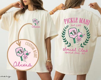 May include: Ivory-colored t-shirt with a front graphic featuring a pickleball paddle, a cocktail, and the name "Olivia." The back of the shirt displays "PICKLE MAHJ est. 2017" and "Social Club" with a laurel wreath design.