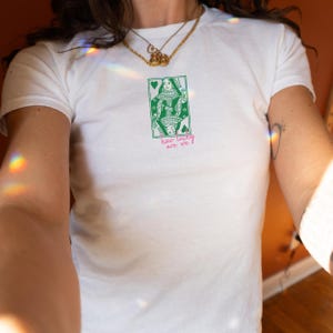 Puede incluir: Camiseta blanca con un dise&ntilde;o de carta de la Reina de Corazones en verde y blanco. El texto "how lucky are we?" est&aacute; impreso en rosa debajo. La camiseta tiene cuello redondo y mangas cortas.