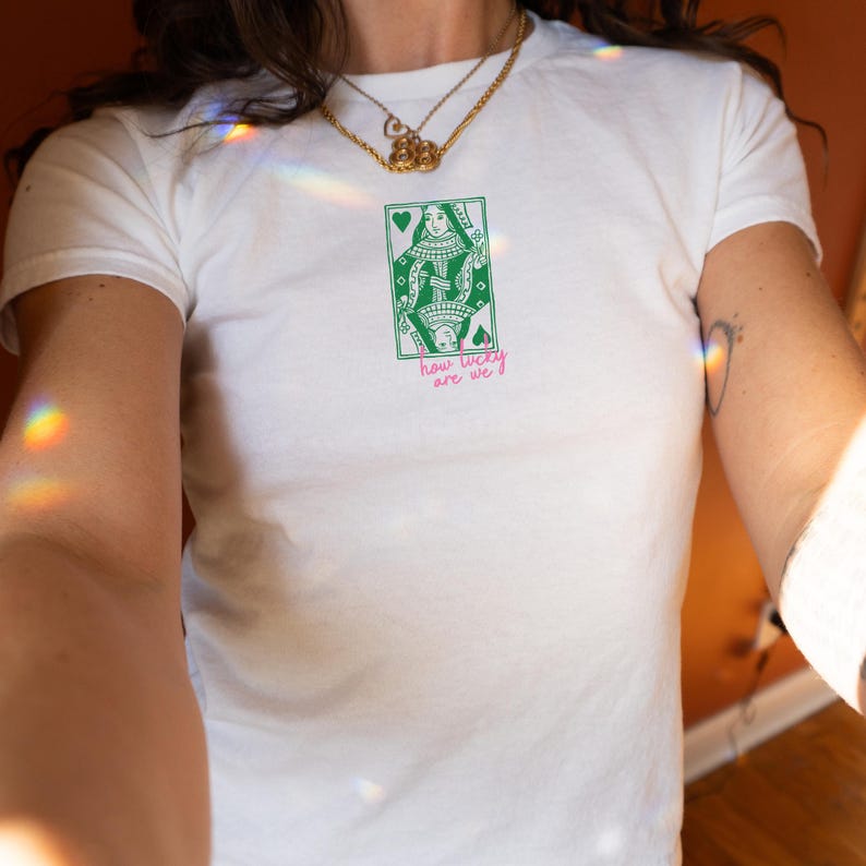 Puede incluir: Camiseta blanca con un dise&ntilde;o de carta de la Reina de Corazones en verde y blanco. El texto "how lucky are we?" est&aacute; impreso en rosa debajo. La camiseta tiene cuello redondo y mangas cortas.