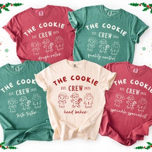 May include: A collection of five t-shirts in various colors, including red, green, and cream. Each shirt features the text "THE COOKIE CREW EST. 2025" with different gingerbread character designs and job titles like "dough roller" and "head baker."