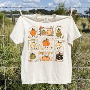 May include: A cream-colored short-sleeve t-shirt featuring a central graphic print of various cartoon pumpkins with smiling faces. The pumpkins are arranged in decorative frames and patterns, including polka dots, plaid, and solid orange designs.