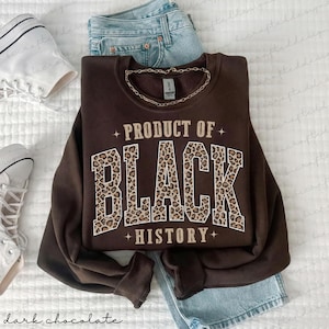 May include: A dark chocolate-colored sweatshirt with the text "PRODUCT OF BLACK HISTORY" in a leopard print design. The sweatshirt is folded on top of a pair of light blue jeans. A silver chain necklace is on the sweatshirt.