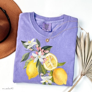 May include: A violet Comfort Colors t-shirt featuring a lemon and floral design. The graphic includes yellow lemons, white flowers, and green leaves. A gold necklace with a pendant rests on the shirt.