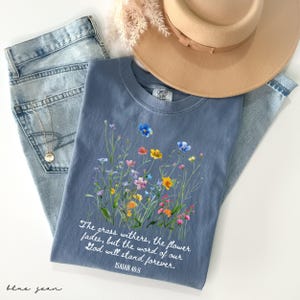 May include: A blue t-shirt with a floral design and the text "The grass withers, the flower fades, but the word of our God will stand forever. ISAIAH 40:8." The shirt is paired with blue jeans and a tan hat.