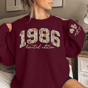Puede incluir: Sudadera burdeos con el a&ntilde;o "1986" en un dise&ntilde;o floral, con las palabras "limited edition" debajo. La manga muestra "Chapter 38" en un coraz&oacute;n floral.
