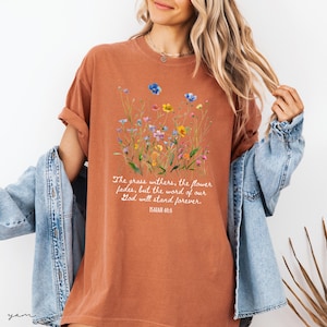 May include: Rust-colored t-shirt featuring a watercolor floral design with blue, yellow, and pink flowers. The text "The grass withers, the flower fades, but the word of our God will stand forever. Isaiah 40:8" is printed below the flowers.