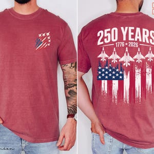May include: A crimson short-sleeve t-shirt featuring a small graphic on the front left chest of fighter jets and a distressed American flag. The back displays the text 250 YEARS 1776-2026 above five fighter jets and a large distressed American flag.
