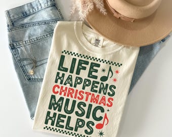 May include: Cream-colored t-shirt with the text "LIFE HAPPENS CHRISTMAS MUSIC HELPS" in green and red. The shirt is paired with light-wash jeans, a tan hat, and a delicate necklace. A sprig of dried flowers adds a touch of elegance.