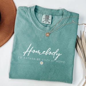 May include: A teal blue t-shirt with the text "Homebody I'd rather be cozy at home." in white lettering with a white daisy graphic.
