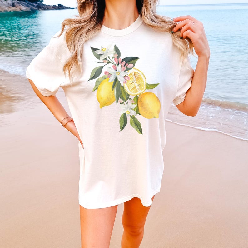 May include: Cream-colored t-shirt featuring a watercolor illustration of lemons, blossoms, and green leaves. The design includes whole and sliced lemons. The shirt has short sleeves and a relaxed fit, suitable for casual wear.