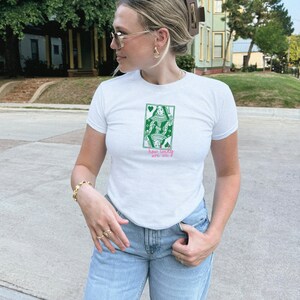 Puede incluir: Camiseta blanca con un dise&ntilde;o gr&aacute;fico verde y rosa de una reina de la baraja. El texto "how lucky are we" est&aacute; debajo. La camiseta se combina con unos vaqueros azul claro.