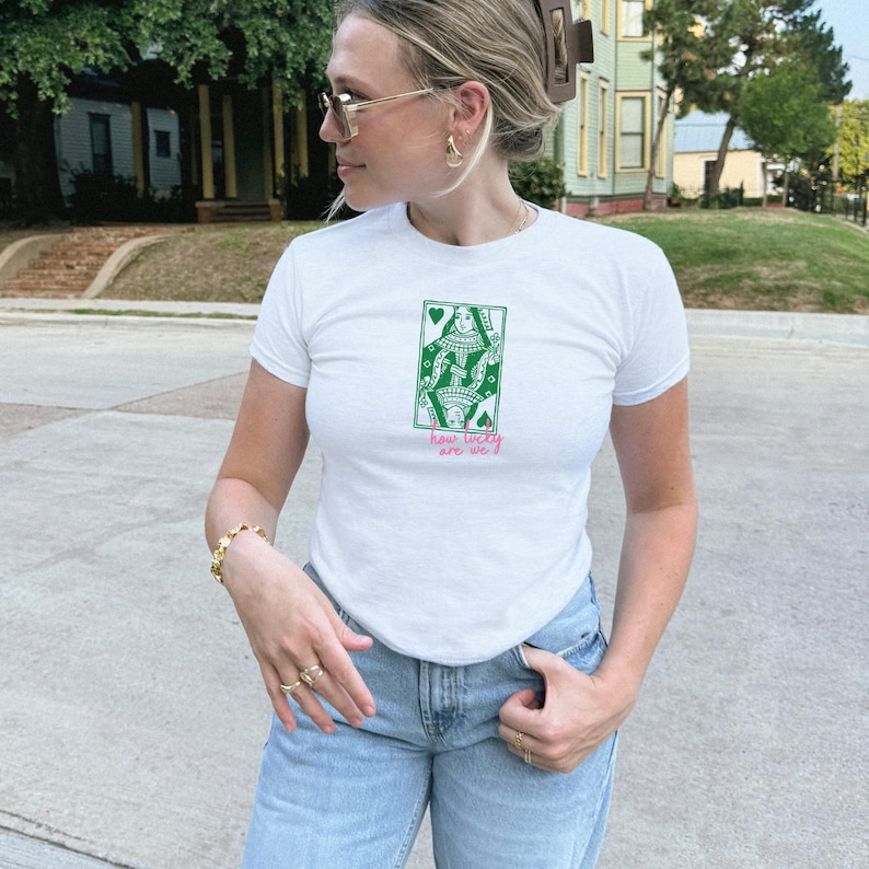 Puede incluir: Camiseta blanca con un dise&ntilde;o gr&aacute;fico verde y rosa de una reina de la baraja. El texto "how lucky are we" est&aacute; debajo. La camiseta se combina con unos vaqueros azul claro.