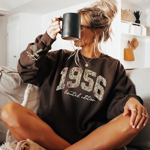 May include: A dark brown sweatshirt with the year "1956" in a floral design. The words "Limited Edition" are below the year. The left sleeve has the words "Vintage Things" printed on it. A black mug is held up.