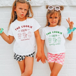 May include: Two young girls wearing white t-shirts with a "The Cookie Crew" graphic and gingerbread person illustrations. One shirt has red text and says "head baker," the other has green text and says "quality control." One girl wears checkered shorts, the other pink checkered shorts.