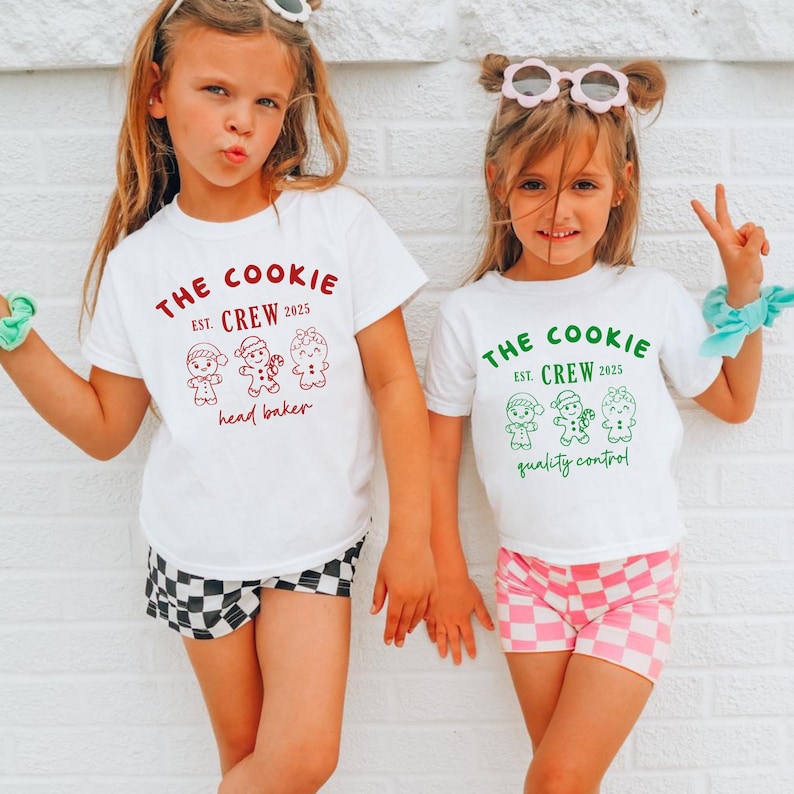 May include: Two young girls wearing white t-shirts with a "The Cookie Crew" graphic and gingerbread person illustrations. One shirt has red text and says "head baker," the other has green text and says "quality control." One girl wears checkered shorts, the other pink checkered shorts.