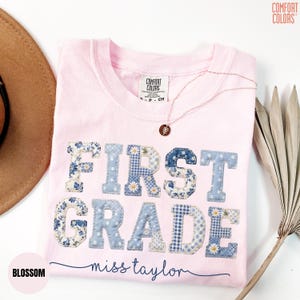 May include: A light pink t-shirt with the words "FIRST GRADE" appliqued in blue and floral patterned fabric. The name "miss taylor" is written in cursive below the text. A gold necklace with a pendant is on the shirt.