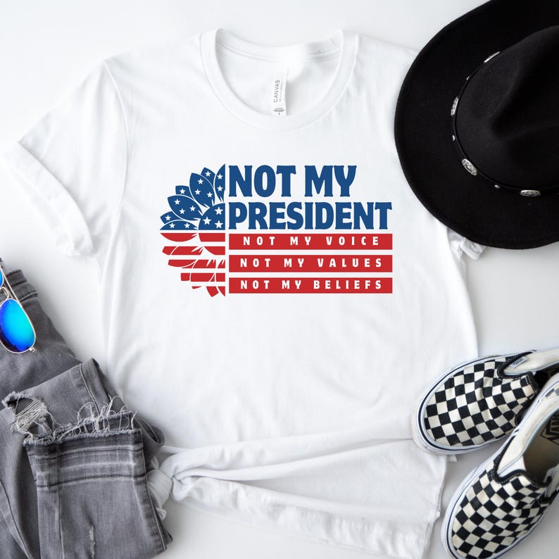 May include: White t-shirt with a red, white, and blue sunflower design. The text "Not My President" is printed in blue, with the words "Not My Voice", "Not My Values", and "Not My Beliefs" printed in black below.