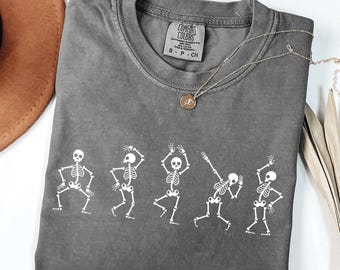 May include: A gray Comfort Colors t-shirt featuring a white graphic of five dancing skeletons. The shirt has a crew neck and short sleeves. A silver necklace with a pendant is draped on the shirt. A brown hat is visible in the background.