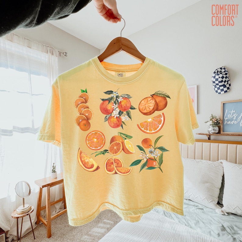 May include: A yellow Comfort Colors t-shirt featuring a vibrant orange fruit design. The graphic includes whole oranges, orange slices, and orange blossoms. The shirt is held up by a wooden hanger.