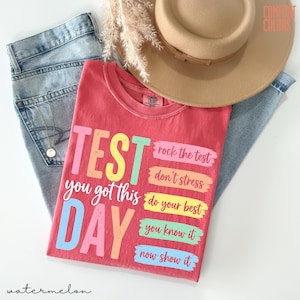 May include: A red t-shirt with the words "TEST DAY" in large, colourful letters. The shirt also has the phrases "you got this," "rock the test," "don't stress," "do your best," "you know it," and "now show it." The shirt is paired with blue jeans and a beige hat.