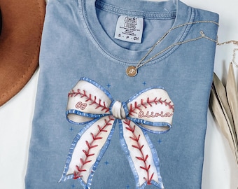 May include: A light blue t-shirt with a white and red baseball-themed bow with the number 88 and the name Olivia. The shirt is made of a soft, comfortable material.