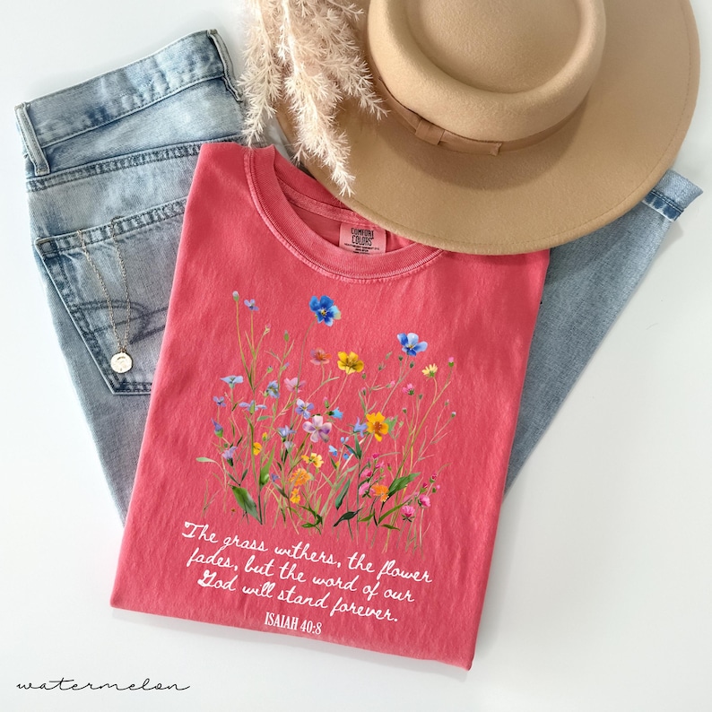 May include: A watermelon-colored t-shirt with a floral design and the text "The grass withers, the flower fades, but the word of our God will stand forever. Isaiah 40:8." The shirt is paired with light blue jeans and a tan hat.