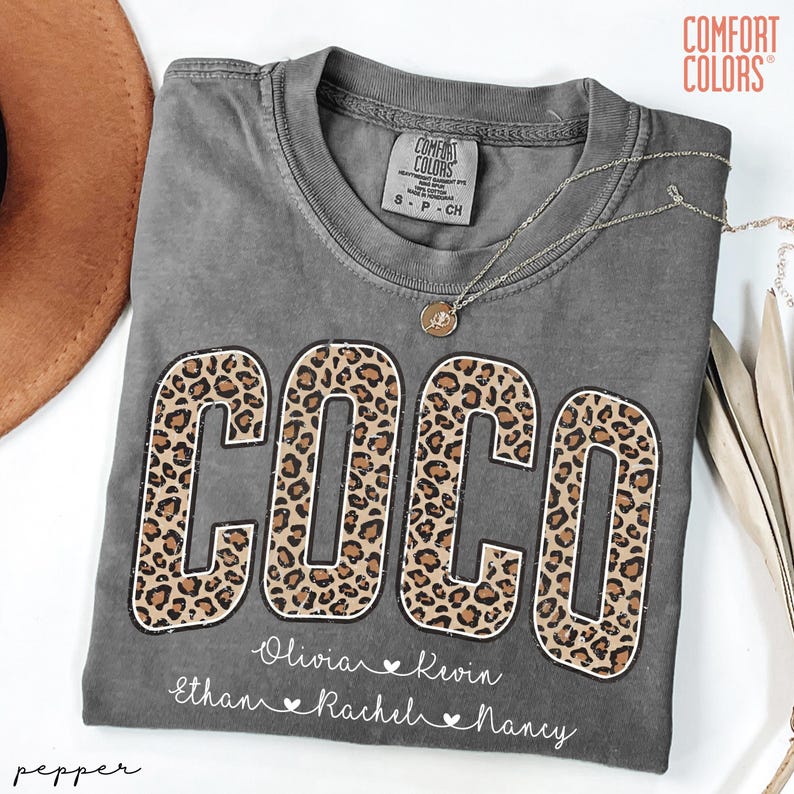 May include: A gray Comfort Colors t-shirt with the name "COCO" in large leopard print letters. Below the name are the names Olivia, Kevin, Ethan, Rachel, and Nancy. A silver necklace with a pendant is draped across the shirt.
