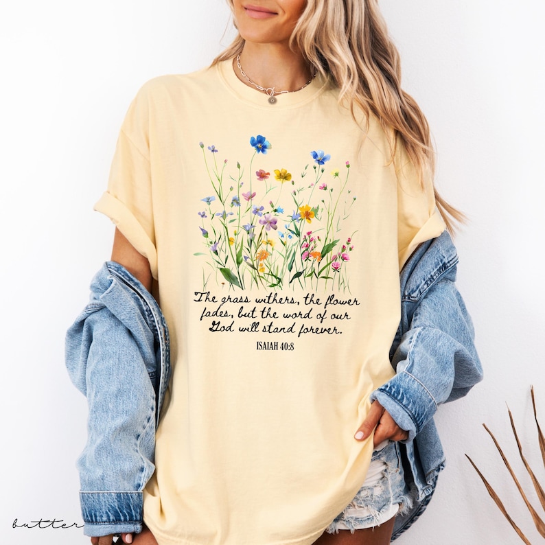 May include: A light yellow t-shirt featuring a floral design with blue, yellow, and purple flowers. The shirt has the text "The grass withers, the flower fades, but the word of our God will stand forever. ISAIAH 40:8."