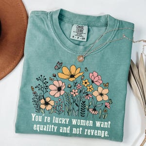 May include: A teal Comfort Colors t-shirt with a floral design featuring yellow and pink flowers and butterflies. The shirt has the text "You're lucky women want equality and not revenge." A silver necklace with a pendant is draped across the shirt.