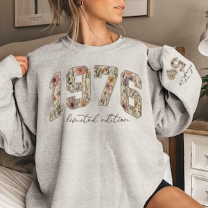 May include: A light gray sweatshirt with the year "1976" in a floral design. The words "limited edition" are below the numbers. The sleeve has the text "50 chapter" with floral hearts.