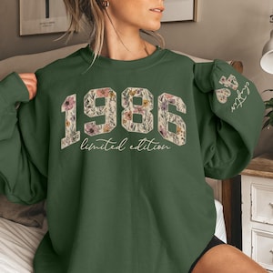 Puede incluir: Una sudadera verde oscuro con el a&ntilde;o "1986" en un dise&ntilde;o floral en la parte delantera. Las palabras "limited edition" est&aacute;n debajo de los n&uacute;meros. La manga tiene el texto "45 chapter" en blanco.