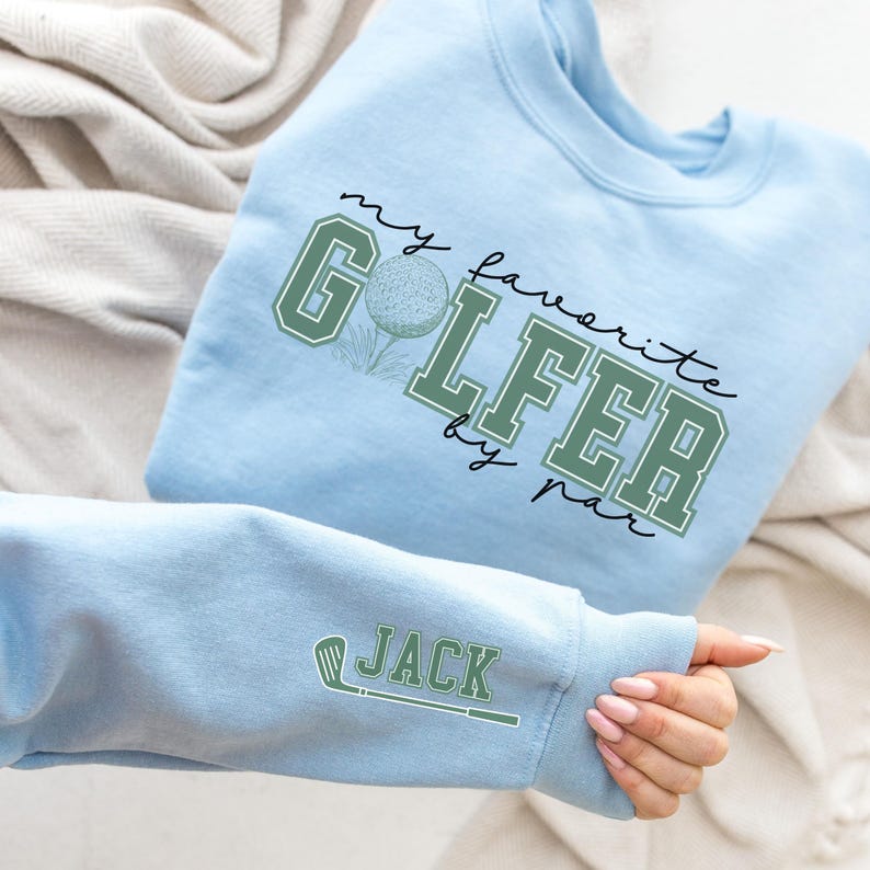 K&ouml;nnte beinhalten: Hellblauer Pullover mit dem Text "my favorite golfer by par" in schwarzer Schrift und einer gr&uuml;nen Golfballgrafik. Der &Auml;rmel hat eine gr&uuml;ne Golfclubgrafik und den Text "JACK" in Schwarz.