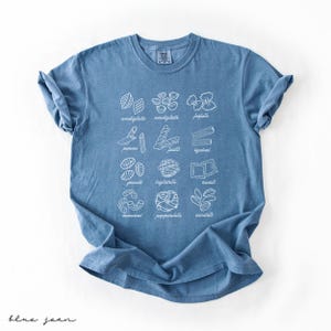 Op de afbeelding: Een blauw jeans T-shirt met een witte afbeelding van verschillende soorten pasta met hun Italiaanse namen. De namen zijn:  conchiglie, farfalle, penne, fusilli, rigatoni, gnocchi, lasagne, ravioli,  macaroni,  pappardelle, en  orecchiette.