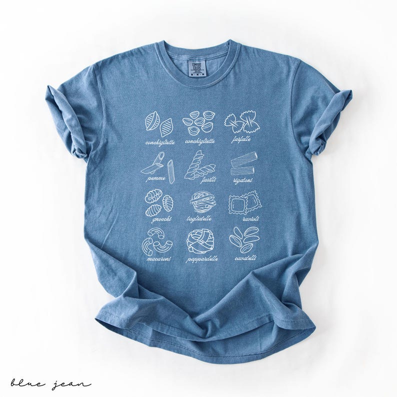 Op de afbeelding: Een blauw jeans T-shirt met een witte afbeelding van verschillende soorten pasta met hun Italiaanse namen. De namen zijn:  conchiglie, farfalle, penne, fusilli, rigatoni, gnocchi, lasagne, ravioli,  macaroni,  pappardelle, en  orecchiette.