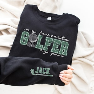 K&ouml;nnte beinhalten: Schwarzer Pullover mit gr&uuml;nem Text, der "my favorite golfer by par" mit einer Golfballgrafik lautet. Der &Auml;rmel hat eine wei&szlig;e Golfclubgrafik und den Text "JACK" in Gr&uuml;n.